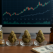 Three jars of cannabis on a wooden desk with a financial graph on a computer screen and a stack of dollar bills.